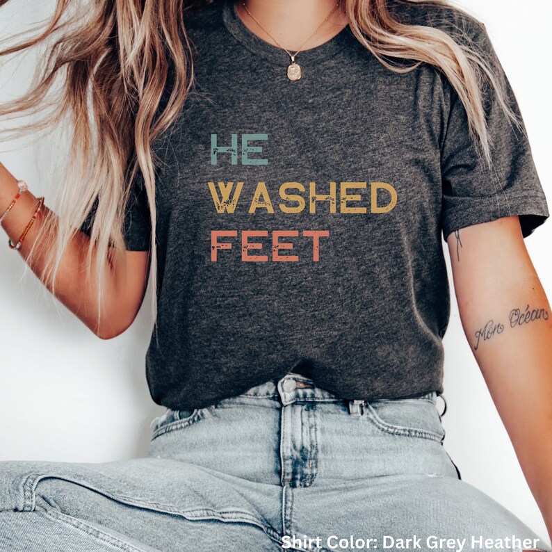 May include: A dark grey heather t-shirt with the text "HE WASHED FEET" in a vintage style font. The text is in different colors, including blue, yellow, and pink.