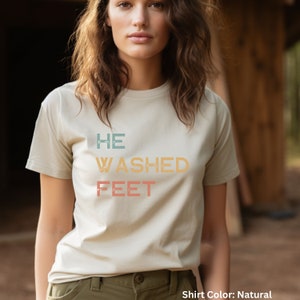 May include: A natural colored t-shirt with the text "HE WASHED FEET" in a vintage style font with a color gradient.