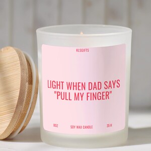 May include: A frosted glass soy wax candle with a light pink label featuring the text KLSGIFTS, LIGHT WHEN DAD SAYS PULL MY FINGER, 8OZ, and 35 H. The candle has a round bamboo lid with a visible wood grain pattern placed to the left of the jar.