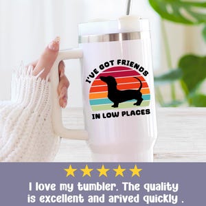 May include: White travel mug with a handle, featuring a black dachshund silhouette against a colorful sunset design. The text reads "I'VE GOT FRIENDS IN LOW PLACES". A straw is inserted in the lid.