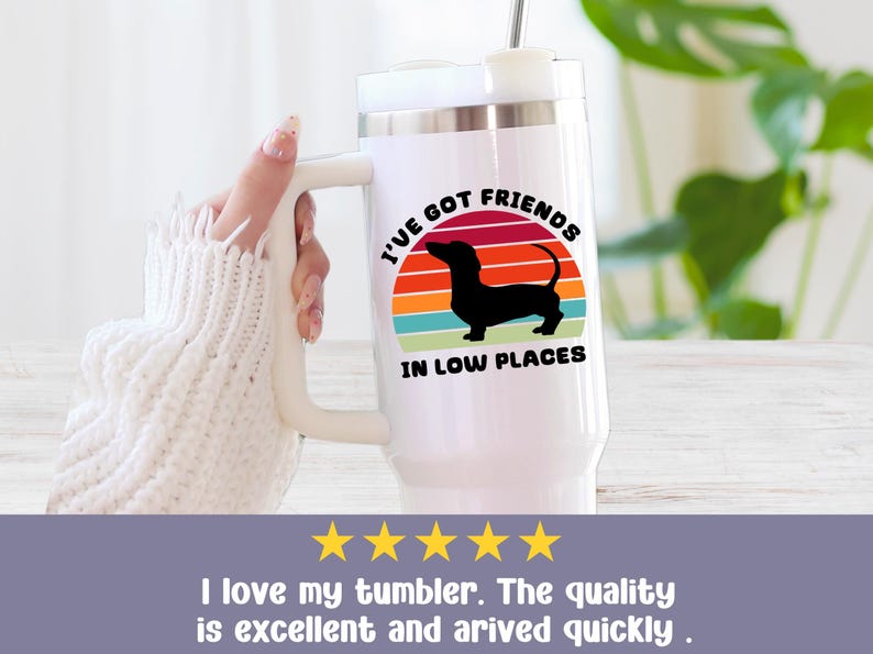 May include: White travel mug with a handle, featuring a black dachshund silhouette against a colorful sunset design. The text reads "I'VE GOT FRIENDS IN LOW PLACES". A straw is inserted in the lid.