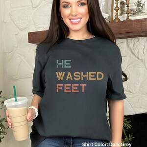 May include: A dark gray t-shirt with the text "HE WASHED FEET" in a vintage style font. The text is in shades of blue, orange, and yellow. Shirt Color: Dark Grey
