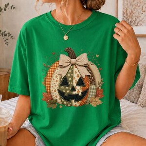 Op de afbeelding: Een felgroen T-shirt met korte mouwen en een grote centrale afbeelding van een pompoen in patchworkstijl. De pompoen is versierd met een beige strik, diverse herfstpatronen en omringd door kleine oranje en bruine herfstbladeren.
