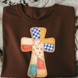 May include: A brown t-shirt featuring a patchwork cross design. The cross is made of various fabric patterns, including floral, gingham, and solid colors. The t-shirt is folded, and the design is centered on the front.