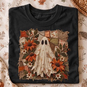 Puede incluir: Una camiseta negra con un diseño de parche cuadrado central. El parche muestra un fantasma blanco rodeado de girasoles naranjas, pequeñas flores blancas y hojas verdes, sobre un fondo de varios cuadrados de tela con texturas.