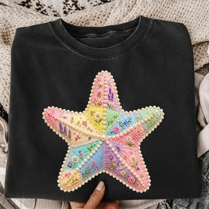 May include: Black crew neck sweatshirt featuring a large, colorful embroidered starfish. The starfish is made of various pastel-colored patches, decorated with floral embroidery and faux pearls. The design is centered on the front of the shirt.