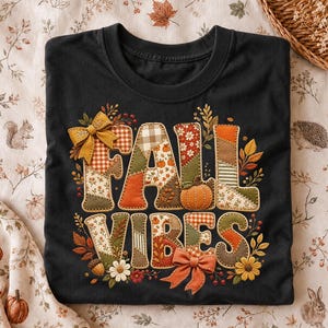 以下が含まれることがあります： パッチワーク風のフォントで「FALL VIBES」という文字がプリントされた黒いTシャツ。文字にはカボチャ、葉、花などの秋らしい模様が施されています。文字には2つのオレンジ色のリボンが付けられ、周囲には花のアクセントが添えられています。