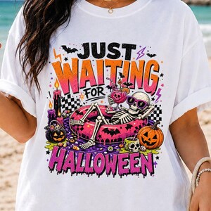May include: A white short-sleeve t-shirt featuring a graphic of a skeleton wearing sunglasses, lounging in a pink inflatable pool float. The design includes the text JUST WAITING FOR HALLOWEEN, pumpkins, candles, bats, and lightning bolts.