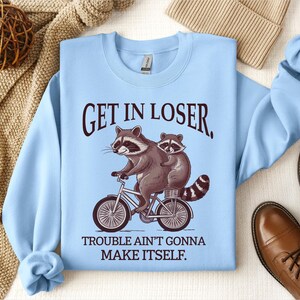 May include: Light blue sweatshirt with the text "GET IN LOSER, TROUBLE AIN'T GONNA MAKE ITSELF." Features a graphic of two raccoons riding a bicycle. The sweatshirt has long sleeves and a crew neck.