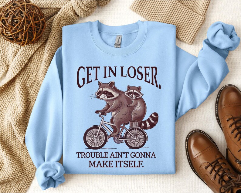 May include: Light blue sweatshirt with the text "GET IN LOSER, TROUBLE AIN'T GONNA MAKE ITSELF." Features a graphic of two raccoons riding a bicycle. The sweatshirt has long sleeves and a crew neck.