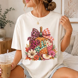 May include: A cream-coloured t-shirt featuring a graphic of a pumpkin decorated with a leopard print bow, a plaid bow, and a patchwork pattern. The pumpkin is surrounded by colourful autumn flowers and leaves, with a slice of apple at the base.