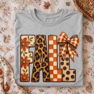 May include: A light grey t-shirt featuring the word FALL in large block letters. Each letter has a unique pattern: the F has orange flowers, the A has a leopard print, the L has an orange checkerboard, and the second L has brown polka dots.