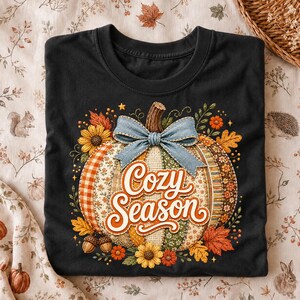 Könnte beinhalten: Ein schwarzes T-Shirt mit einer großen Grafik eines Kürbisses, der mit verschiedenen Stoffmustern, einer blauen Schleife im Jeans-Look und dem Schriftzug "Cozy Season" in weißer Schreibschrift verziert ist. Herbstblätter, Sonnenblumen und Eicheln umgeben ihn.