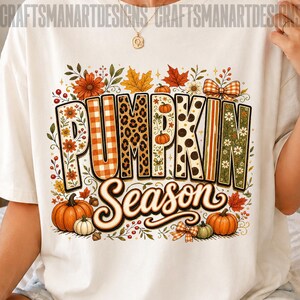 May include: A white t-shirt featuring a graphic print with the text "PUMPKIN Season". The letters are filled with various patterns including orange gingham, leopard print, and floral designs. The graphic includes pumpkins, autumn leaves, and bows.