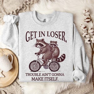 May include: A light gray sweatshirt with a raccoon riding a bicycle graphic. The raccoon is brown and gray with a black and white striped tail. The text "GET IN LOSER, TROUBLE AIN'T GONNA MAKE ITSELF" is printed above and below the graphic.