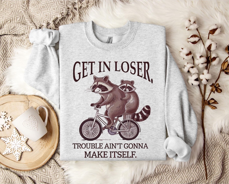 May include: A light gray sweatshirt with a raccoon riding a bicycle graphic. The raccoon is brown and gray with a black and white striped tail. The text "GET IN LOSER, TROUBLE AIN'T GONNA MAKE ITSELF" is printed above and below the graphic.