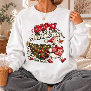 May include: A white crewneck sweatshirt featuring a graphic of a white goose wearing a red and white patterned sweater, hat, and boots. The goose is carrying a decorated Christmas tree. The text reads OOPS! I GOT ANOTHER TREE.