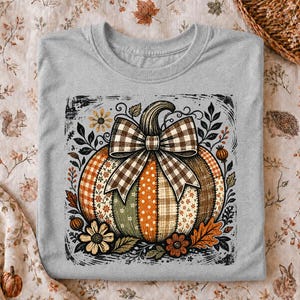 May include: A heather grey t-shirt featuring a central graphic of a pumpkin composed of various patterned segments in orange, green, and brown. The pumpkin is adorned with a large brown and white gingham bow and surrounded by floral motifs.
