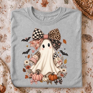 Op de afbeelding: Een grijs gemêleerd t-shirt met een centrale afbeelding van een witte geest die een grote strik draagt met luipaardprint, ruitjes en bloemenpatronen. De geest is omringd door kleine zwarte vleermuizen, oranje en roze pompoenen en bloemen.