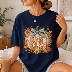 Könnte beinhalten: Ein marineblaues T-Shirt mit einer zentralen Grafik eines Kürbisses, der mit verschiedenen herbstlichen Mustern, einer hellblauen Schleife und dem Schriftzug "Fall Vibes" in Schreibschrift verziert ist, umgeben von Sonnenblumen und Herbstblättern.