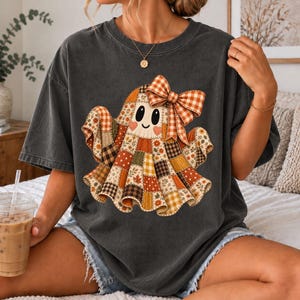 Op de afbeelding: Een antracietgrijs t-shirt met korte mouwen en een centrale afbeelding van een geest gemaakt van diverse herfstachtige patchworkpatronen in oranje, bruine en crèmekleuren, versierd met een grote oranje en wit geblokte strik op het hoofd.