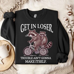 May include: Black sweatshirt with the text "GET IN LOSER." A cartoon raccoon rides a bicycle with another raccoon on the back. The text "TROUBLE AIN'T GONNA MAKE ITSELF" is below the image. The sweatshirt has long sleeves.