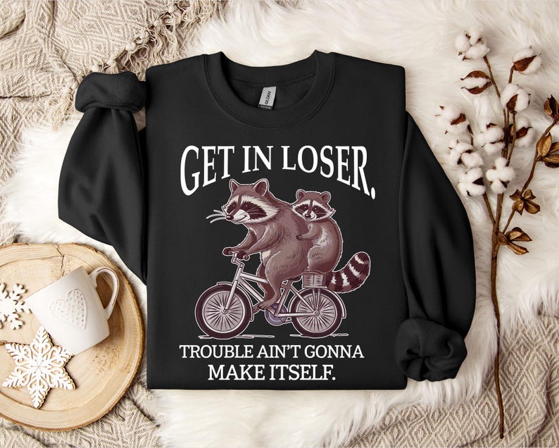 May include: Black sweatshirt with the text "GET IN LOSER." A cartoon raccoon rides a bicycle with another raccoon on the back. The text "TROUBLE AIN'T GONNA MAKE ITSELF" is below the image. The sweatshirt has long sleeves.