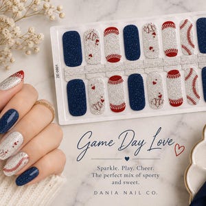 May include: A sheet of adhesive nail wraps featuring a baseball theme with navy blue glitter, silver glitter, and red stitching patterns. Some wraps include small red heart accents. The text on the sheet reads Game Day Love, Sparkle. Play. Cheer. The perfect mix of sporty and sweet. DANIA NAIL CO.