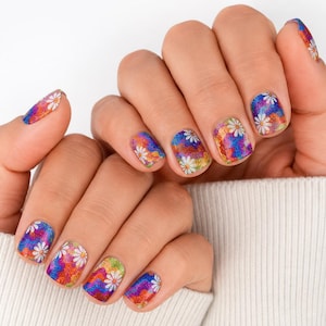 May include: Nail wraps featuring a vibrant, multi-colored wavy pattern in shades of blue, purple, orange, and green with a glittery finish. The design is accented with small, white daisy flower motifs scattered across each fingernail.