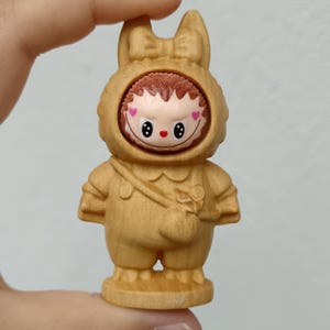 Peut inclure: Figurine en bois d'un personnage en costume de lapin. Le personnage a un visage souriant avec des yeux noirs et des joues roses en forme de cœur. Le costume est marron clair avec un petit sac et un nœud.