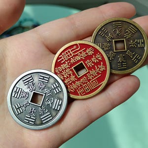 Op de afbeelding: Drie antieke Chinese munten met vierkante gaten in het midden worden getoond. De munten zijn in zilver, rood en brons, elk met ingewikkelde reliëftekens. De munten worden in een hand gehouden, wat hun gedetailleerde ontwerp en historische esthetiek laat zien.