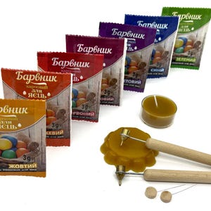 May include: Assortment of six food coloring packets for dyeing Easter eggs. Each packet contains 3 grams of coloring and is labeled with the color name in Russian. The colors are red, orange, yellow, green, blue, and purple. The packets are arranged in a row from left to right, with the red packet at the left and the purple packet at the right. There is also a small candle and two wooden tools for decorating eggs.