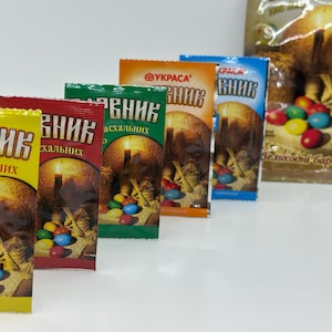 Puede incluir: Seis paquetes de tinte para huevos de Pascua en varios colores, incluyendo amarillo, rojo, verde y azul. Cada paquete presenta una vela y una cesta de huevos de Pascua. El texto en los paquetes dice "Красник Пасхальних", que se traduce como "Tinte de Pascua".