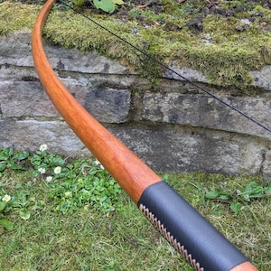 Könnte beinhalten: Ein Holzbogen mit einem schwarzen Ledergriff und braunen Nähten. Der Bogen liegt auf einem Bett aus grünem Gras.