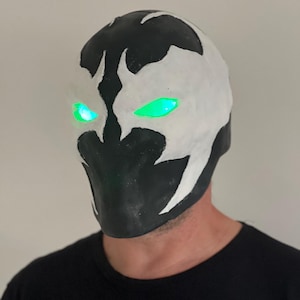 Puede incluir: Una máscara en blanco y negro con ojos verdes brillantes. La máscara tiene un diseño distintivo con una cara blanca y detalles en negro. Los ojos brillan intensamente, creando un efecto visual llamativo. La máscara es probablemente para cosplay o disfraces.