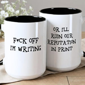 May include: Two white ceramic mugs with black interiors and handles. One mug reads "F*ck Off I'm Writing" in black text. The other mug says "Or I'll Ruin Our Reputation In Print". The mugs are on a light brown surface.