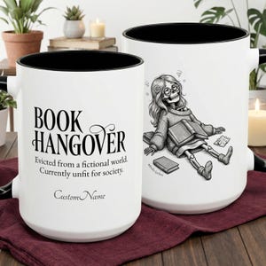 Puede incluir: Dos tazas de cerámica blancas con bordes y asas negras. Una taza tiene el texto "BOOK HANGOVER" y la otra una ilustración de esqueleto. El texto dice: "Expulsado de un mundo ficticio. Actualmente no apto para la sociedad."