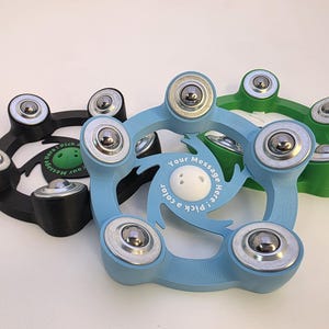 May include: Three colorful fidget spinners in black, light blue, and green. Each spinner has a circular design with six silver bearings and a central white disc. Text on the spinners reads "Your Message Here! Pick a color".