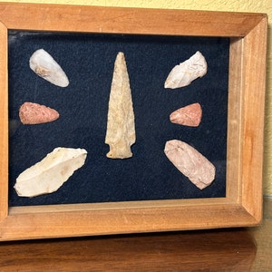 May include: A framed display of eight arrowheads, including a large, pointed arrowhead in the center. The arrowheads are arranged in a symmetrical pattern on a dark blue background.