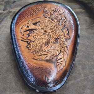 May include: A brown leather motorcycle seat with an embossed eagle design. The seat has a dark border and a textured pattern around the edges. The eagle's head and feathers are intricately carved, showing fine detail.