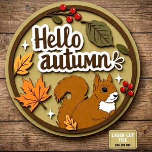 Op de afbeelding: Een rond, gelaagd houten bord met de tekst Hello autumn in bruin handschrift. Het ontwerp bevat een bruine eekhoorn, oranje esdoornbladeren, groene bladeren en rode bessen op een licht olijfgroene achtergrond. Een vakje toont LASER CUT FILE.