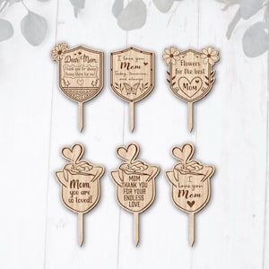 Puede incluir: Decoraciones de madera para el Día de la Madre con mensajes como "Querida mamá" y "Te amo mamá". Las decoraciones tienen forma de escudos y diseños florales, ideales para celebrar a las madres.