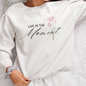 May include: White sweatshirt with the text "LIVE IN THE Moment" in black script and a watercolor floral design. The floral design features pink lotus flowers and green stems. The sweatshirt is part of a matching set with white sweatpants.