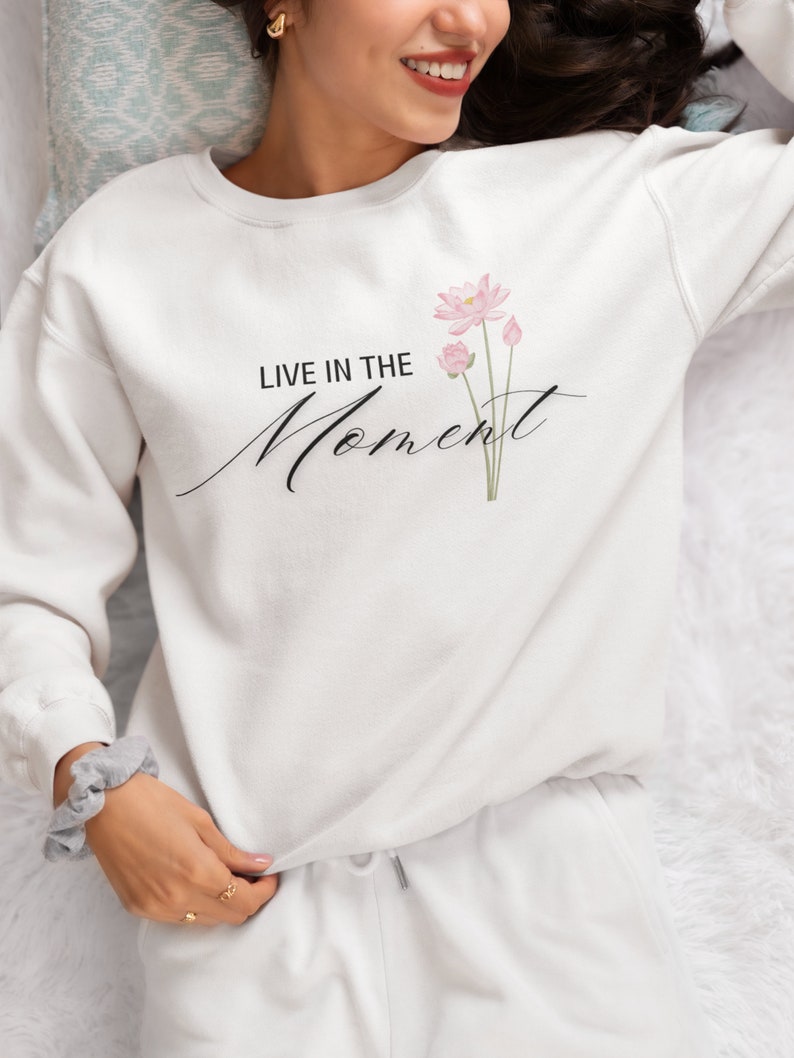 May include: White sweatshirt with the text "LIVE IN THE Moment" in black script and a watercolor floral design. The floral design features pink lotus flowers and green stems. The sweatshirt is part of a matching set with white sweatpants.