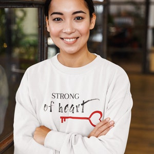 Puede incluir: Una sudadera color crema con las palabras "STRONG of heart" impresas en negro y un diseño de llave roja. El mango de la llave tiene forma de corazón. La persona que lleva la sudadera está sonriendo.