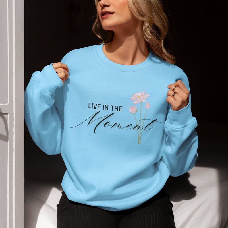 May include: Light blue sweatshirt with the text "LIVE IN THE Moment" in black script and a floral design. The floral design features pink flowers with green stems. The sweatshirt is a crew neck style.
