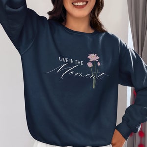 May include: Navy blue sweatshirt with the text "LIVE IN THE Moment" in a cursive font. A small floral design with pink flowers and green stems is printed on the front. The sweatshirt is paired with white pants.