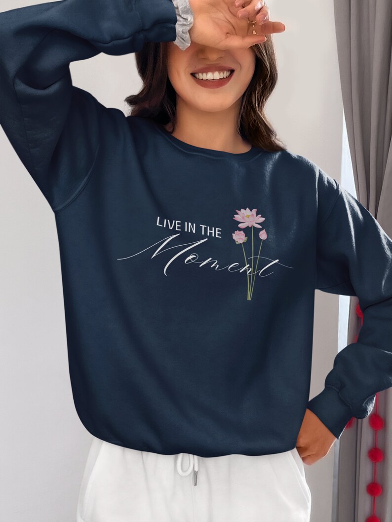 May include: Navy blue sweatshirt with the text "LIVE IN THE Moment" in a cursive font. A small floral design with pink flowers and green stems is printed on the front. The sweatshirt is paired with white pants.