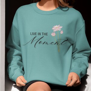 May include: Teal-colored sweatshirt with the text "LIVE IN THE Moment" in a cursive font. A floral design of pink lotus flowers and buds is printed on the right side of the text. The sweatshirt is a solid color and has a crew neck.