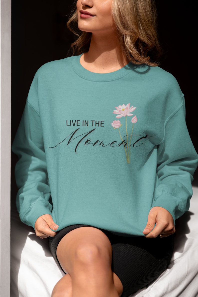 May include: Teal-colored sweatshirt with the text "LIVE IN THE Moment" in a cursive font. A floral design of pink lotus flowers and buds is printed on the right side of the text. The sweatshirt is a solid color and has a crew neck.